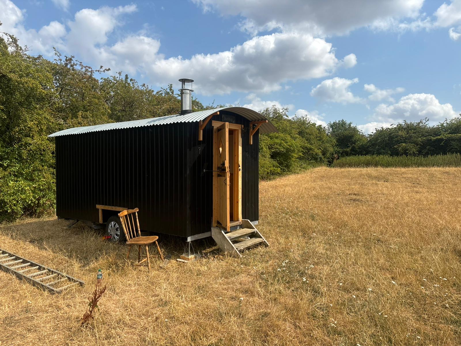 A black-clad shepherd's hut named Buddug in a sunny hay meadow, with its door open and a chair set outside.