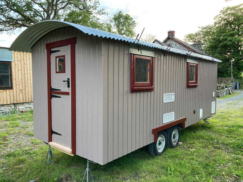 A mocha-toned shepherd's hut with dark red trim, on its trailer wheels in evening light.
