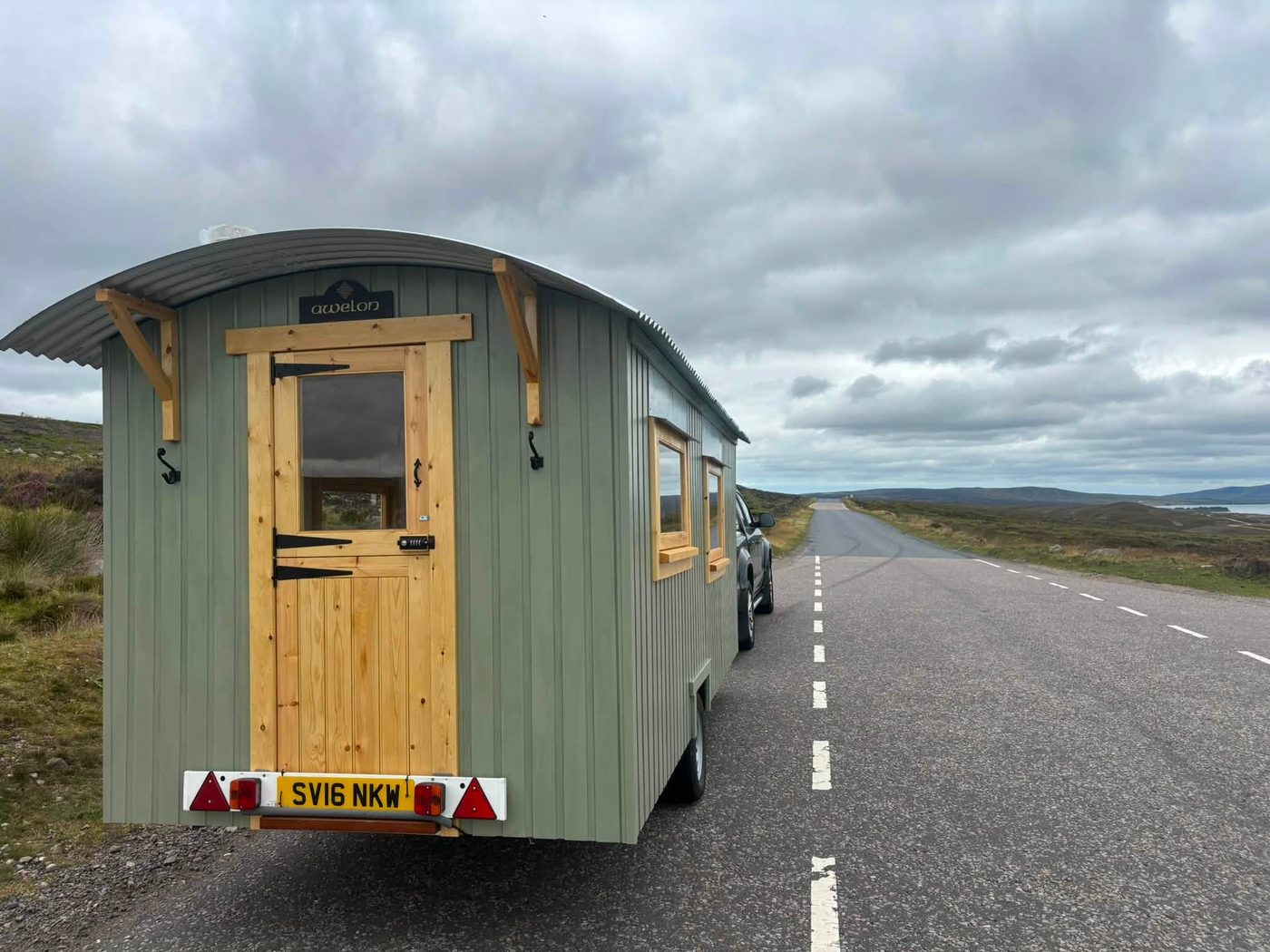 A sage green shepherd hut being towed along a Welsh moorland road.