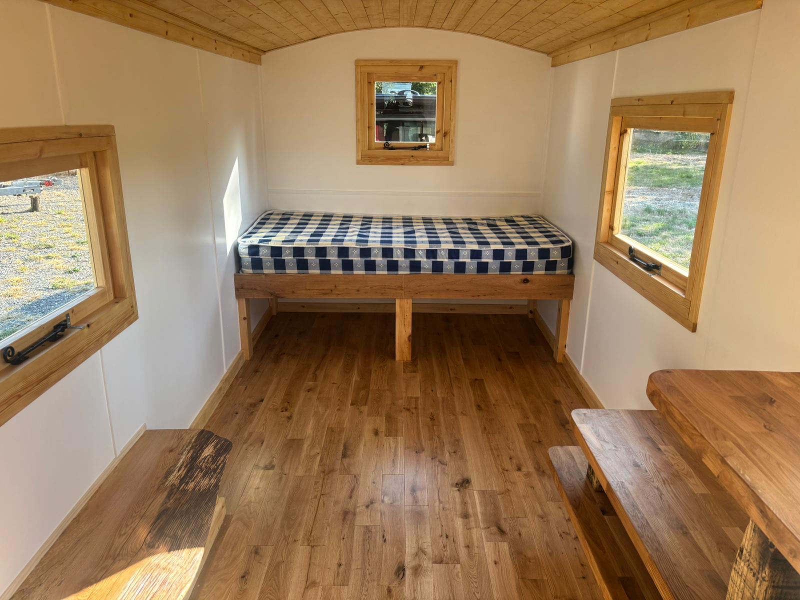 A single bed with a blue check quilt, lit by windows on both sides of the hut.
