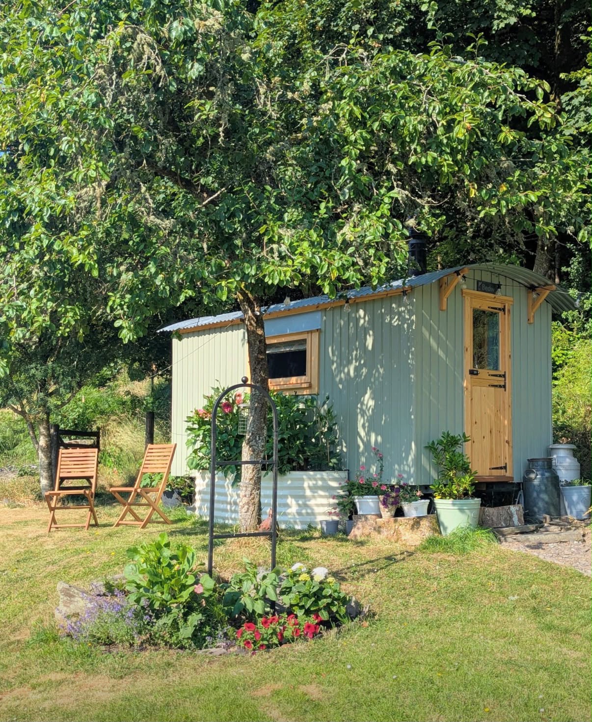 A sage green shepherd\'s hut with a timber stable door, under an apple tree with flowers and garden chairs.
