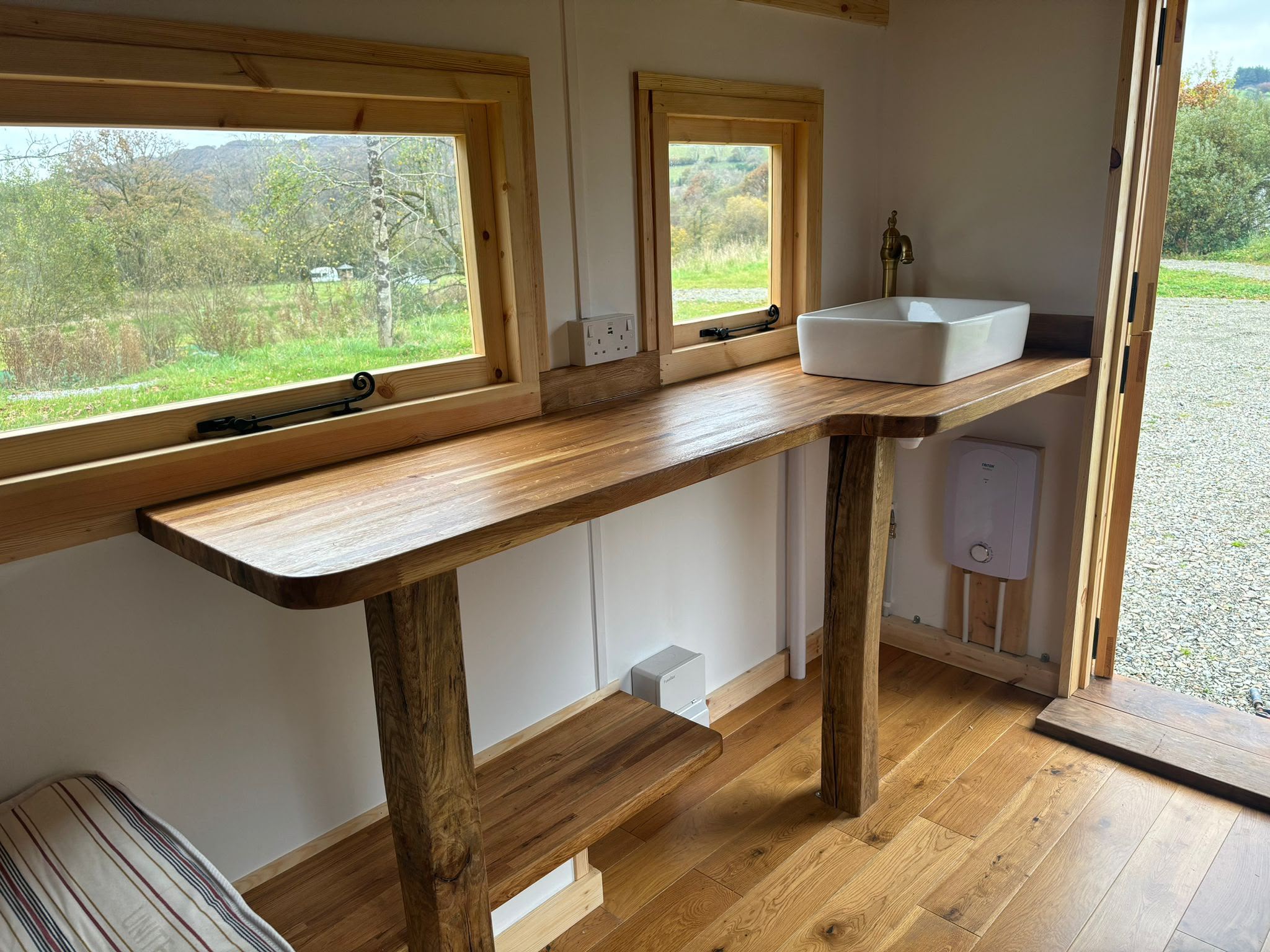Oak worktop with a ceramic basin and brass tap inside a shepherd hut, windows looking onto fields.